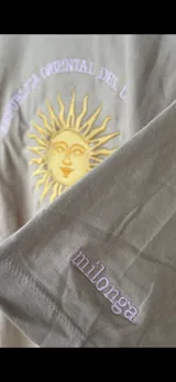 Remera de manga corta y cuello redondo, color beige claro, con un bordado central que representa el Sol de Mayo con el texto "REPUBLICA ORIENTAL DEL URUGUAY" y "EST. 1825".