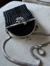 Cartera de mano tejida a crochet color negro, con cierre metálico plateado y cadena larga delgada. Presenta aplique de flor con strass en el frente.