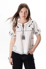 Blusa blanca de mangas cortas con bordados negros de animales y motivos geométricos. Tiene cuello redondo con cordones y borlas decorativas.