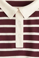 Remera tipo polo de manga larga con corte holgado, a rayas horizontales color borgoña y crema. Tiene cuello clásico color crema, tapeta con botones y hombros caídos.