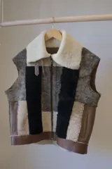 Chaleco sin mangas confeccionado con retazos de cuero en tonos marrón, beige, blanco y negro, con cuello de piel sintética color crema y cierre frontal con cremallera.