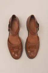 Zapato marrón de cuero vacuno trenzado a mano, con correa al tobillo con hebilla negra y suela de cuero.