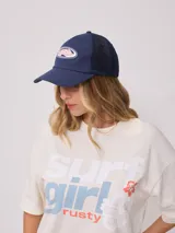 Gorra de lona blanca con visera azul marino y malla trasera azul marino. Presenta un logo bordado en el frente con letras rosadas y contorno azul marino.