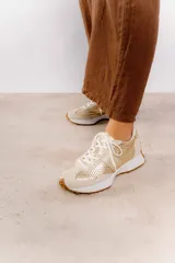 Zapatillas deportivas color beige con detalles metalizados dorados, confeccionadas en cuero con perforaciones, cordones ajustables y suela de goma dentada.