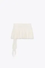 Falda pantalón negra de tiro bajo con cintura drapeada y bajo acabado en volantes superpuestos. Presenta un lazo largo y fino que cae desde el lateral.