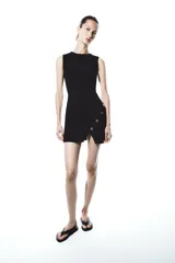 Vestido corto negro de punto, con cuello redondo y sin mangas. Tiene una abertura en el ruedo y detalle de botones metálicos irregulares cruzados en el lateral.