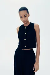 Chaleco cropped negro en punto bouclé con cuello redondo, pinzas en el busto, botones a presión dorados y forro interno.