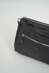 Bandolera color beige, con forma rectangular y extremos redondeados. Tiene una correa ajustable con hebilla negra y cierre de cremallera en la parte superior.