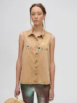 Camisa sin mangas de denim liviano color camel, con cuello camisero y botones frontales. Presenta bordados temáticos marinos en el pecho, incluyendo un sol, olas, una gaviota y vida marina.