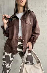 Sobrecamisa marrón de gamuza sintética, corte oversize, con cuello camisero, cierre frontal con botones a presión, mangas largas con puños abotonados, bolsillos de parche en el pecho y detalle de calado con motivos florales en el ruedo.