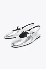 Zapato tipo bailarina destalonada plateada con efecto metalizado, punta redonda y tira delantera con aplique metálico.