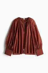 Blusa color ladrillo de tejido brillante de mezcla de lyocell. Modelo oversized con hombros caídos, cuello redondo con cordón para ajustar, abertura en V y tiras finas largas con dijes de cuentas brillantes. Mangas globo largas con costuras raglán en delantero y espalda, y puños vueltos con abertura y botón.