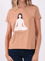 Remera de manga corta color marrón claro con cuello redondo. Estampado frontal de una figura femenina en posición de loto de yoga, con texto debajo que dice "Yoga is not a work-out, it is a work-in".