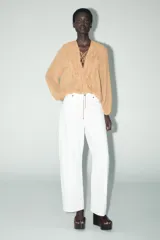 Camisa color marrón claro con escote en pico ajustable con lazada y manga larga abullonada. Detalle de volantes en el mismo tejido en el frente y en el cuello.