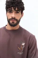 Buzo de algodón color marrón oscuro (ciruela/ciruela quemado) con cuello redondo, puños y ruedo acanalados. Presenta un pequeño bordado de motivos florales en el pecho.