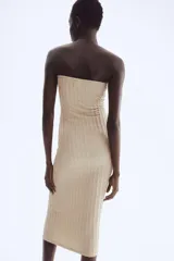 Vestido midi ajustado color beige, sin tirantes (strapless), con escote corazón que incluye un alambre de metal revestido para soporte y un borde de silicona oculto para mejor ajuste. El tejido presenta una textura plisada o arrugada vertical.