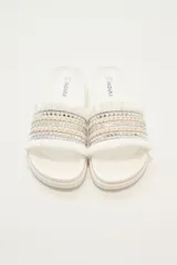 Sandalias tipo slider blancas, con tira ancha decorada con trenzado de hilo beige y plateado, tachas piramidales plateadas y flecos blancos en los bordes.