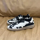 Zapatillas bajas con estampado de vaca en blanco y negro, cordones negros y suela color caramelo.