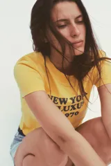 Camiseta amarilla de algodón con cuello redondo, manga corta y estampado marrón con el texto "New York Fluxus 1970".