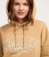 Buzo color tostado de algodón y poliéster, con capucha ajustable con cordón, mangas largas con puños ajustados y bolsillo canguro. Estampa frontal con el texto "Colorado Denver" en color blanco.