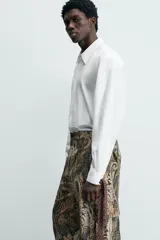 Camisa blanca de algodón popelín, corte relajado, con cuello solapa y pechera frontal plisada. Las mangas terminan en puños ajustables con lazos largos estampados en color marrón con patrón circular.