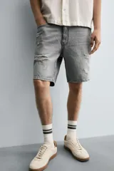 Bermuda de denim de algodón, color gris lavado, con corte relaxed fit y roturas visibles en la parte delantera de ambas piernas. Presenta cinco bolsillos y cierre frontal con cremallera y botón.