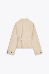 Trench corto color crema con cuello solapa, manga larga con trabilla, bolsillos de doble vivo, forro interior y cinturón lazada en el mismo tejido.