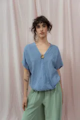 Blusa de lino color azul claro con escote en V cruzado y manga corta. Se ajusta en el centro del busto con un botón redondo de madera o metal con relieve.