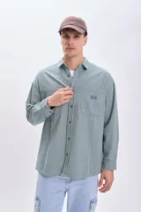 Sobrecamisa verde con rayas verticales blancas, de corte holgado, manga larga, cuello camisero y dos bolsillos frontales con solapa.