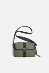 Bandolera color verde militar con acabado engomado. Tiene un compartimento principal con cierre de cremallera y un bolsillo interior con cremallera. En la parte frontal tiene un bolsillo con cierre mediante hebilla de liberación rápida. El asa de hombro es ajustable y extraíble.