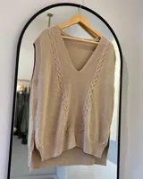Chaleco de hilo color beige con escote en pico, punto elástico en mangas y bajo, detalle de punto elástico y punto en ocho en la delantera, y espalda más larga que la delantera.