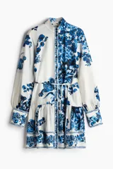 Vestido corto camisero blanco con estampado floral azul, cuello mao, botonadura frontal y cinturón fino para atar extraíble. Mangas globo de corte raglán, puños abotonados y costura fruncida bajo la cadera.
