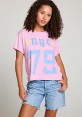 Remera rosa con estampado frontal de las letras "NYC" en azul y el número "79" en azul con borde rosa. Tiene dos rayos amarillos a los costados de las letras.