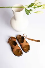 Sandalia plana color camel con tiras cruzadas sobre el empeine y tira para ajuste al tobillo con hebilla. Presenta un diseño de dedo (toe-loop).