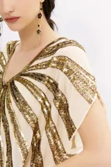 Blusa color crema con escote en V y mangas cortas tipo kimono. Presenta un diseño de rayas horizontales con lentejuelas doradas.