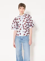 Camisa celeste de poplin con cuello mao, lazo para atar en el cuello, mangas cortas abullonadas y bordado de lentejuelas con forma de flores rojas.