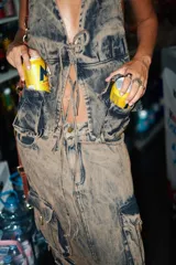 Chaleco tipo top de denim con efecto ahumado (lavado ácido/tie-dye en tonos azules y morados). Presenta escote marcado, bolsillos tipo cargo frontales con argollas metálicas en color bronce y cordones ajustables en el frente.