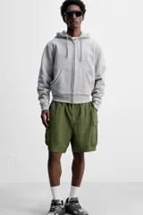 Bermuda color verde militar, relaxed fit, confeccionada en tejido técnico ligero. Cintura elástica ajustable con cordón interior, pliegues frontales, bolsillos delanteros y traseros con solapa, bolsillos cargo con solapa en perneras. Cierre frontal con cremallera y botón.