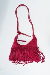Bolso de hombro tipo hobo color rojo oscuro, confeccionado en material tipo gamuza o nobuk, con flecos largos en la parte inferior. La correa es delgada y se anuda en la parte superior.