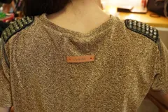 Vestido corto color dorado confeccionado en lycra brillante, con cuello redondo y mangas cortas. Presenta hombreras con tachas y cadenas decorativas.