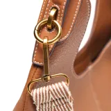 Bolso tipo hobo de cuero sintético (PU) color marrón habano, con textura granulada. Presenta costuras verticales en el centro y un logo metálico XL en la parte inferior frontal. Incluye un asa corta fija y una correa larga desmontable y regulable con diseño tejido a rayas blancas y negras.