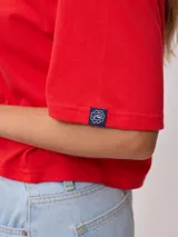 Remera gris de algodón con corte cropped y calce holgado. Tiene cuello redondo y mangas cortas con costuras reforzadas en color coral. Presenta un pequeño logo de la marca Rusty en el pecho.