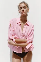Camisa de lino a rayas verticales rosas y blancas, con cuello solapa, manga corta abullonada y lazos en los puños. Cierre frontal con botones ocultos.