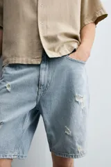 Bermuda de denim de algodón, color gris lavado, con corte relaxed fit y roturas visibles en la parte delantera de ambas piernas. Presenta cinco bolsillos y cierre frontal con cremallera y botón.