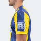 Camiseta de fútbol Le Coq Sportif del Rosario Central, modelo titular temporada 25/26. Diseño a rayas verticales amarillas y azules, con un patrón geométrico sutil en las franjas azules. Presenta cuello en V y detalles en azul en mangas y hombros.