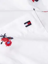 Camisa blanca de manga corta con cuello camisero, cierre de botones frontal y bolsillo de parche en el pecho. Presenta un bordado floral integral en tonos rojo, azul y negro.