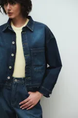 Chaqueta de jean azul oscuro con cuello clásico, cierre frontal con botones metálicos, un bolsillo de parche en el pecho y detalle de lavado en puños y bajo.