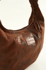 Cartera tipo hobo de gamuza color marrón con etiqueta de cuero con el logo de la marca Foglia.