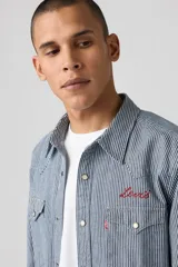 Camisa de manga larga a rayas verticales azules y blancas, con cuello americano, cierre frontal con botones a presión, dos bolsillos con solapa abotonada en el pecho y logo bordado en rojo sobre el bolsillo derecho.