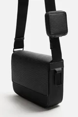 Bandolera negra con acabado engomado y textura acanalada. Tiene un compartimento principal con cierre de solapa y botón a presión, un bolsillo interior con cremallera y un monedero cuadrado en la correa. La correa de hombro es ajustable.